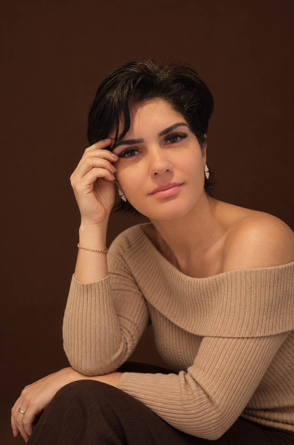 Joice sentada em estúdio com fundo marrom, blusa bege e pulseira dourada.
